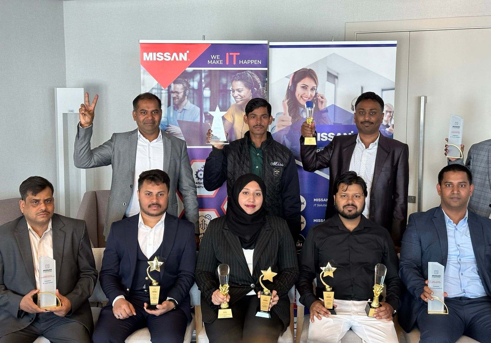 About nine Missan staff holding award trophies, seated and standing in front of Missan banners