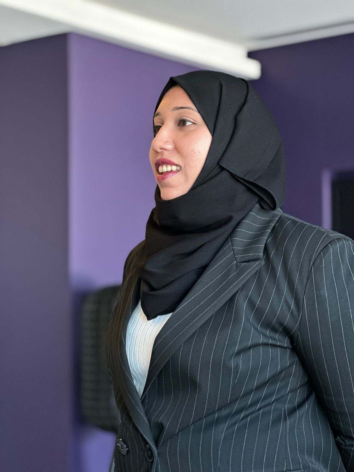 A Missan team member in a pinstripe suit and hijab speaking, seen in profile against a purple wall