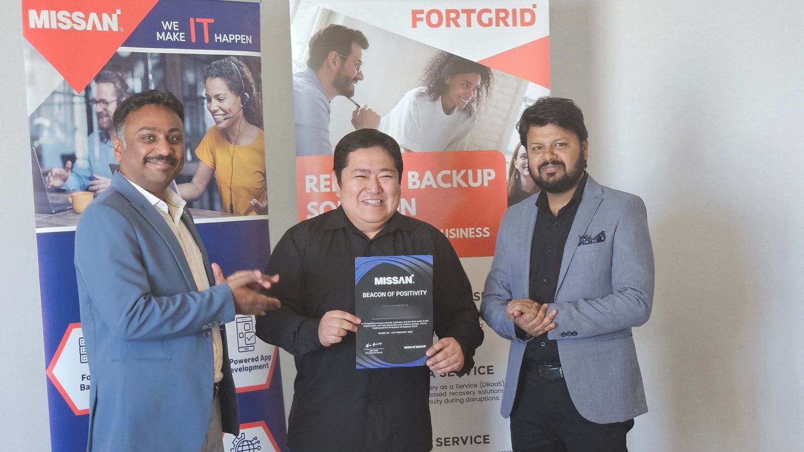 Three Missan staff smiling, the one in the middle holding a certificate, in front of Missan and FortGrid banners