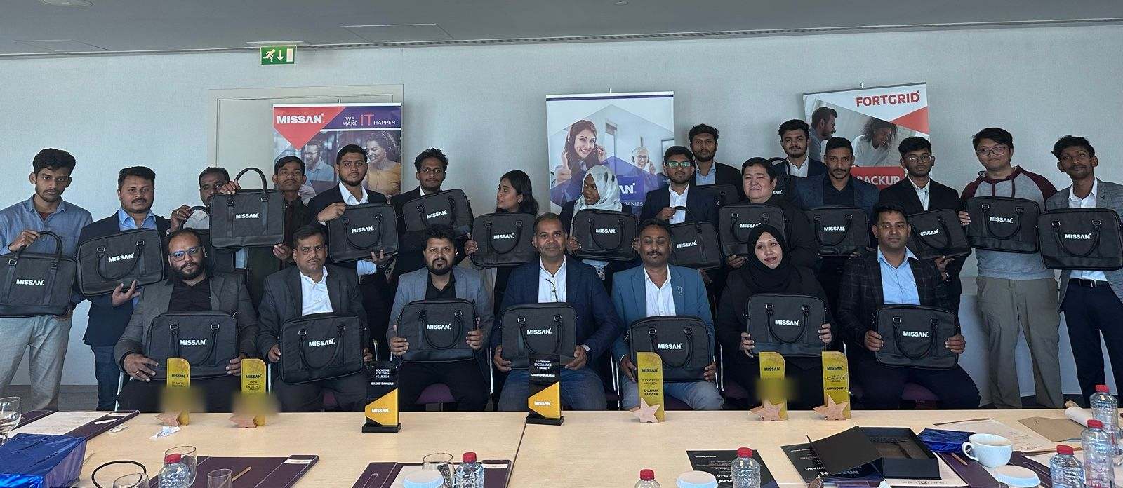 About twenty-four Missan UAE staff in two rows holding Missan-branded bags, with Missan and FortGrid banners behind and award trophies on the table in front