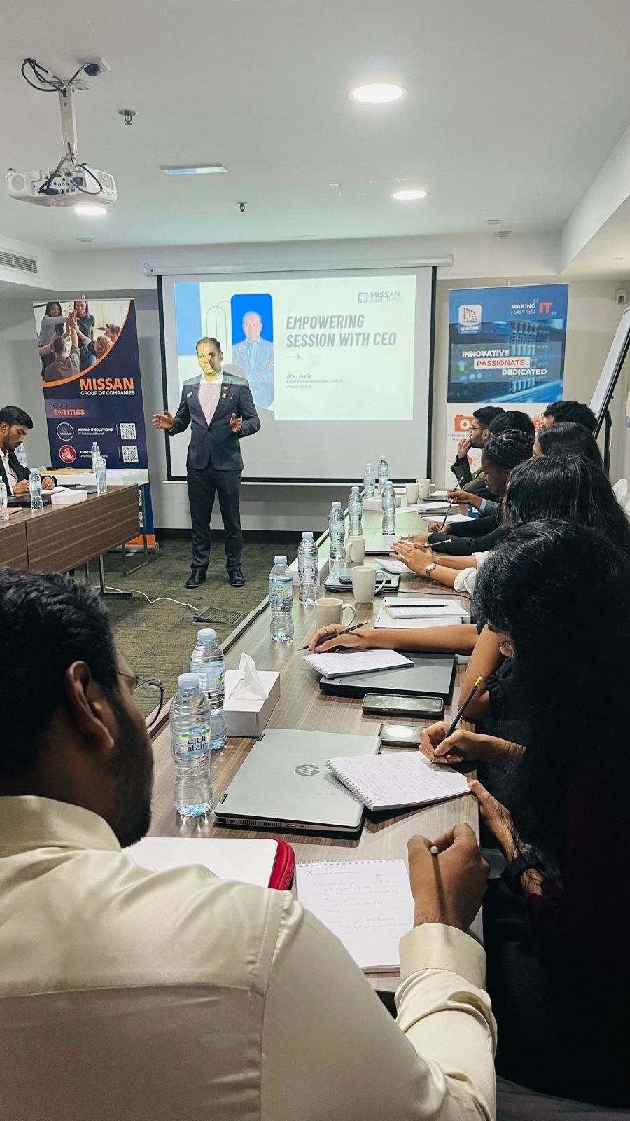 Missan's CEO presenting at the front of a meeting room to attendees seated along a table, taking notes