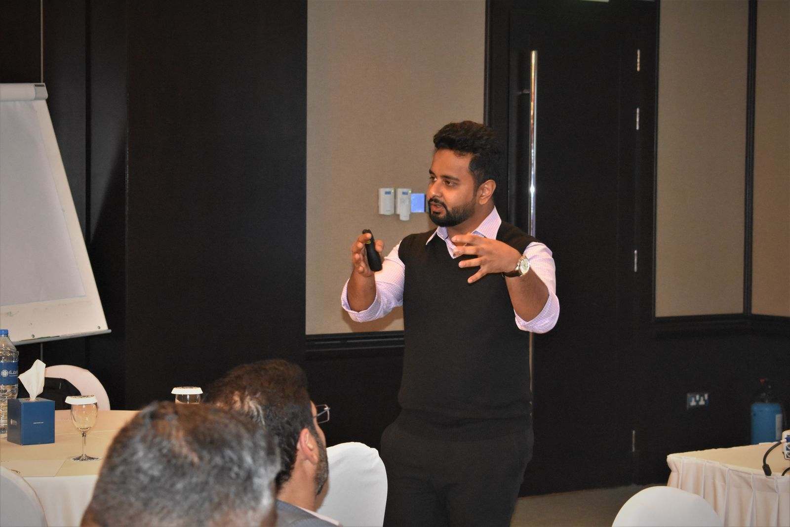 A presenter gesturing as he speaks to an audience seated at round tables