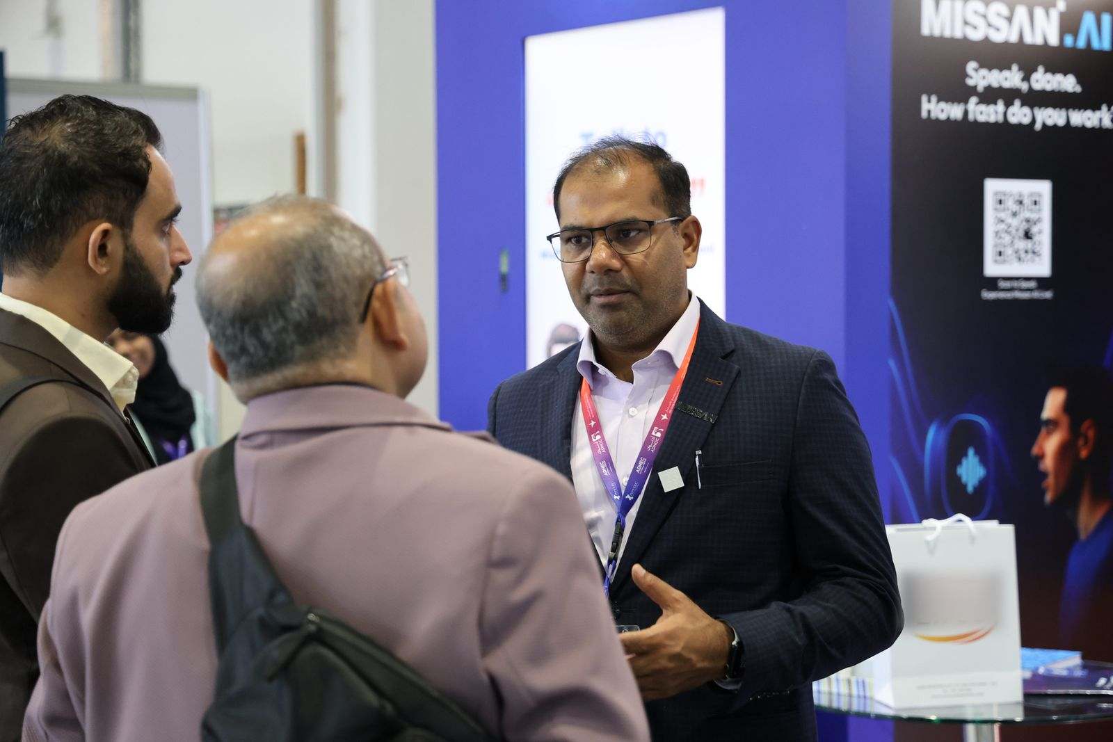 A Missan team member in a dark suit talking with two visitors, seen from behind, in front of the Missan.AI panel