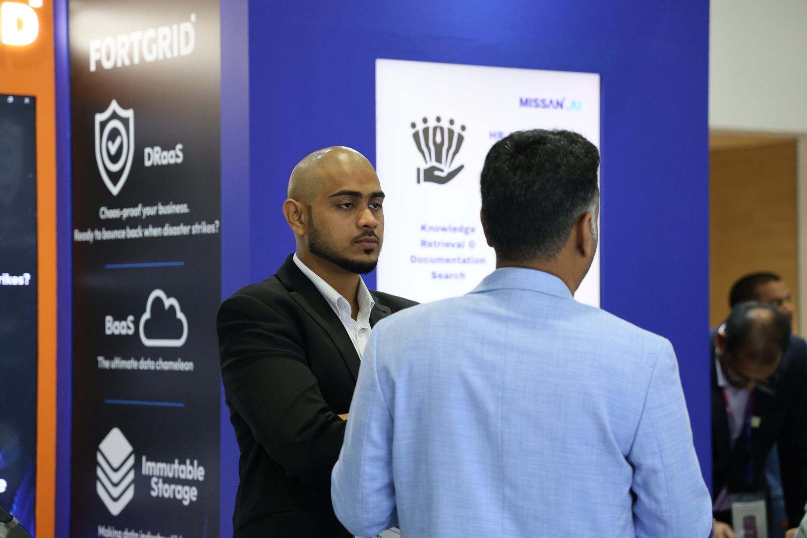 A Missan team member listening to a visitor in a light-blue blazer beside the FortGrid panel