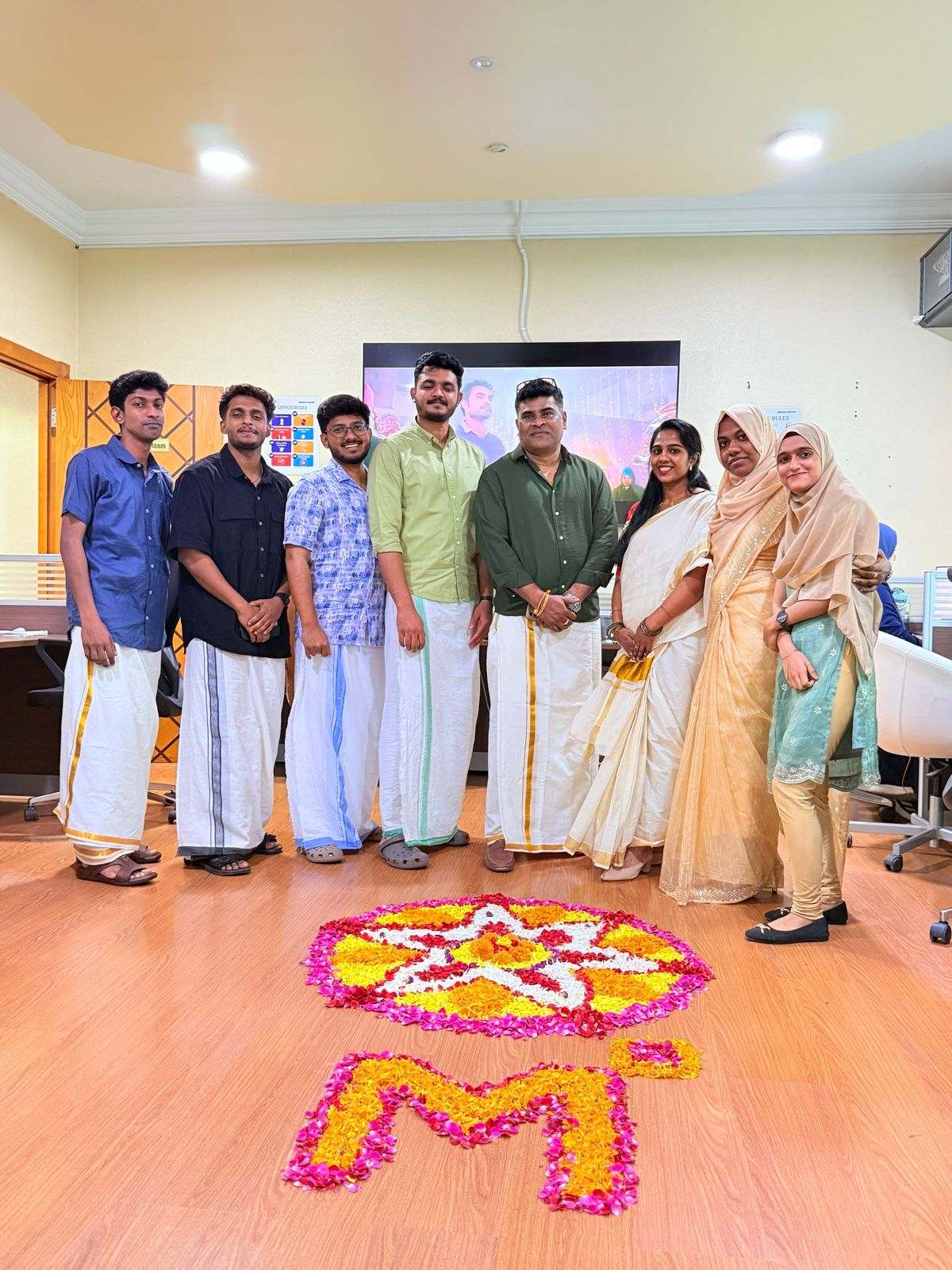 Eight Missan staff in traditional Kerala dress standing behind a flower pookalam that includes a large M