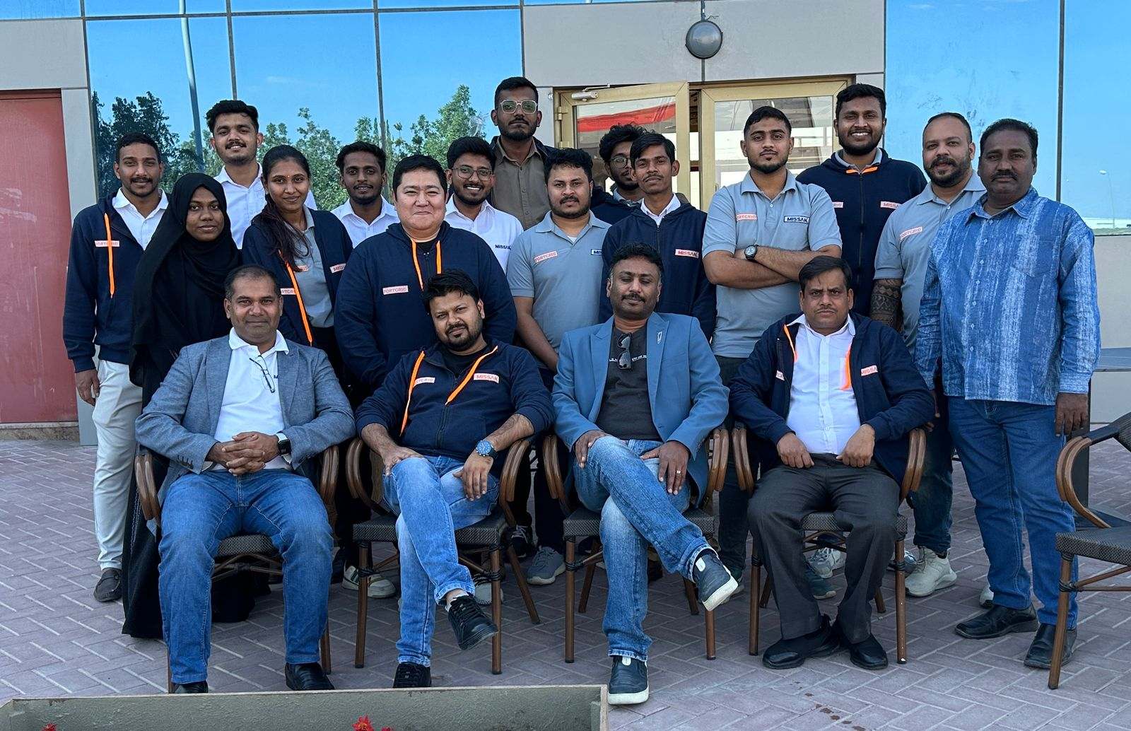 About twenty Missan staff, many in Missan-branded jackets and polo shirts, seated and standing in two rows outside a restaurant